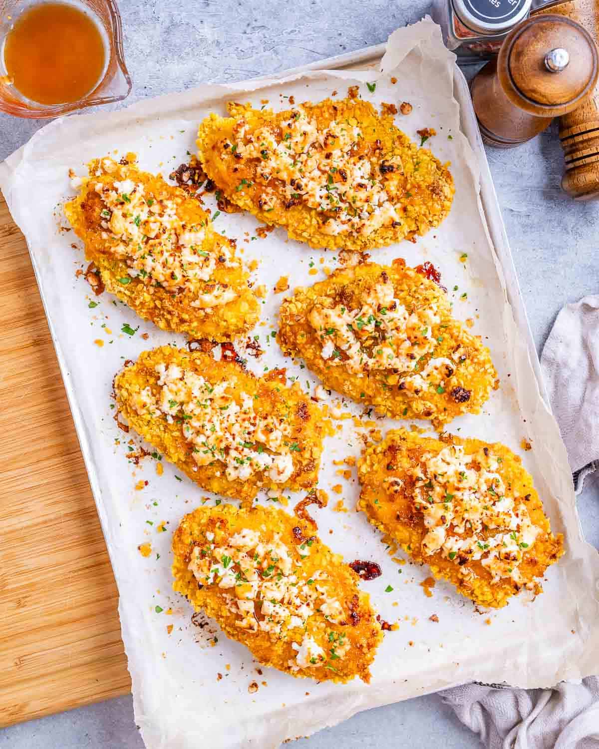 Overhead view of multiple completed chicken cutlets on parchment, topped with feta, hot honey glaze, and scattered chopped green herbs.