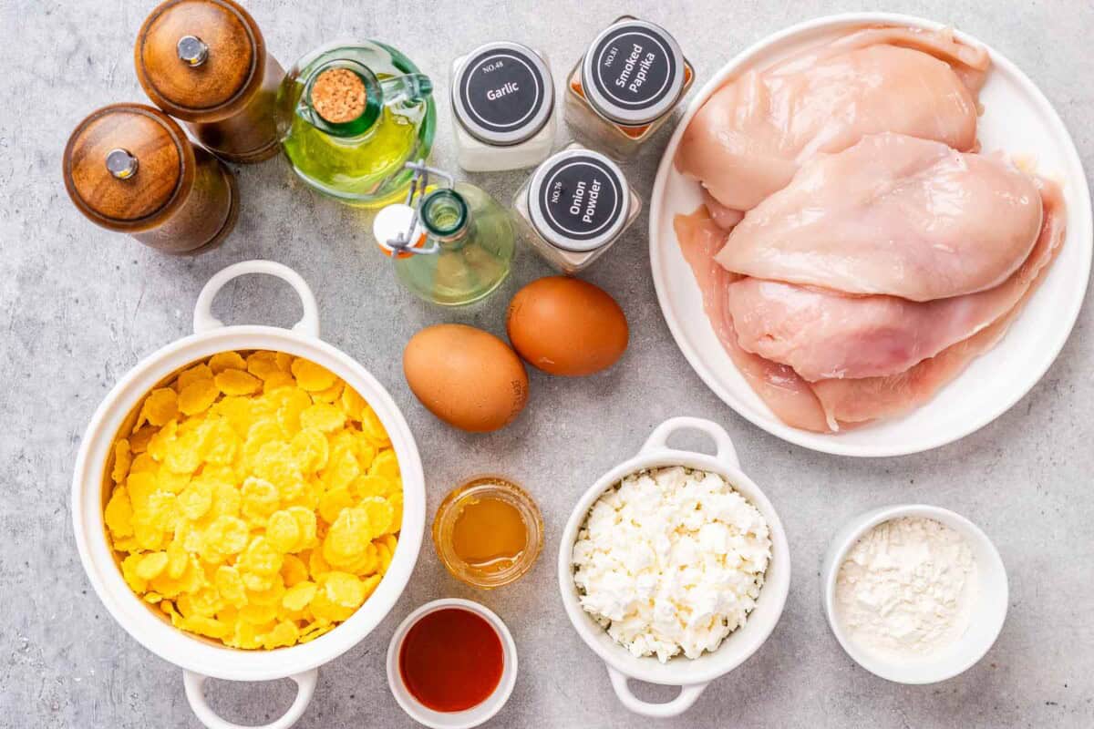Flat lay of recipe ingredients on a countertop: raw chicken cutlets, bowl of beaten eggs, cornflakes, crumbled feta, honey, sriracha, spices, olive oil, and small bowls of seasonings.
