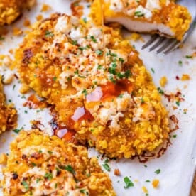 Close up of a a piece of hot honey chicken on parchment, topped with feta, hot honey glaze, and scattered chopped chives.