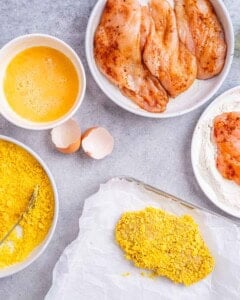 Overhead view of a white bowl filled with beaten eggs, surrounded by cracked eggshells, raw seasoned chicken cutlets, and a piece of chicken covered in cornflakes on a baking tray on parchment paper, all on a light countertop.