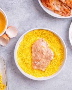 A raw chicken cutlet coated in seasoned crushed cornflakes inside a large white bowl of yellow cornflake mixture.