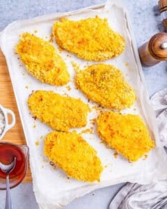 Several breaded chicken cutlets arranged in a single layer on a parchment-lined baking sheet, showing thick golden cornflake coating.