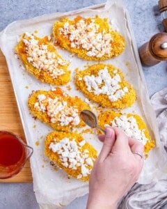 Hand drizzling hot honey sauce over the white feta cheese on top of hot golden baked chicken cutlets on a parchment-lined sheet pan.