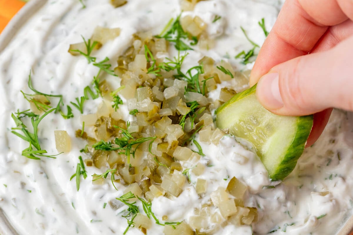 a hand dipping cucumber into the dill pickle yogurt dip.