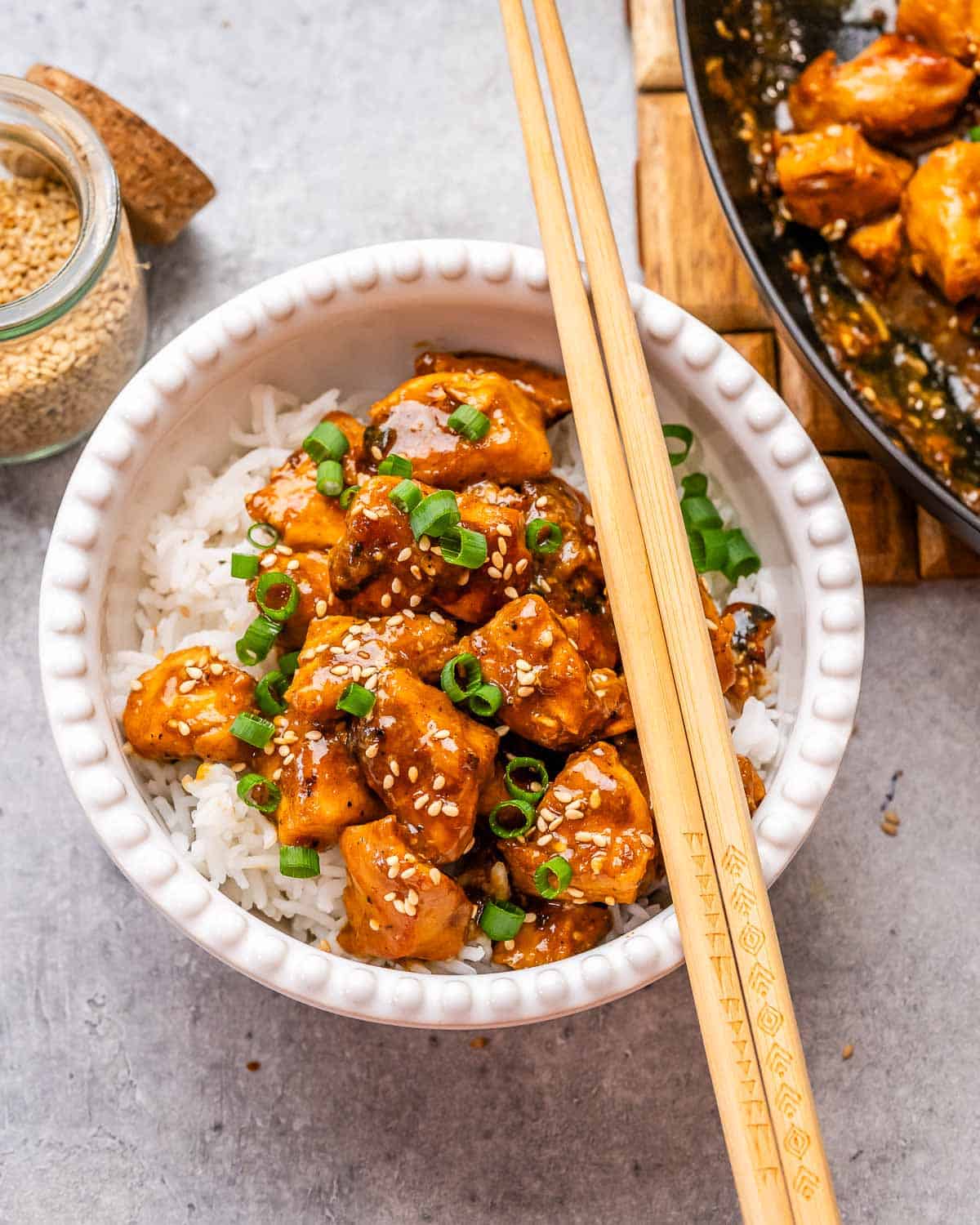 A white bowl with hosin chicken and rice, garnished with scallions and sesame seeds, with chopsticks on the side.