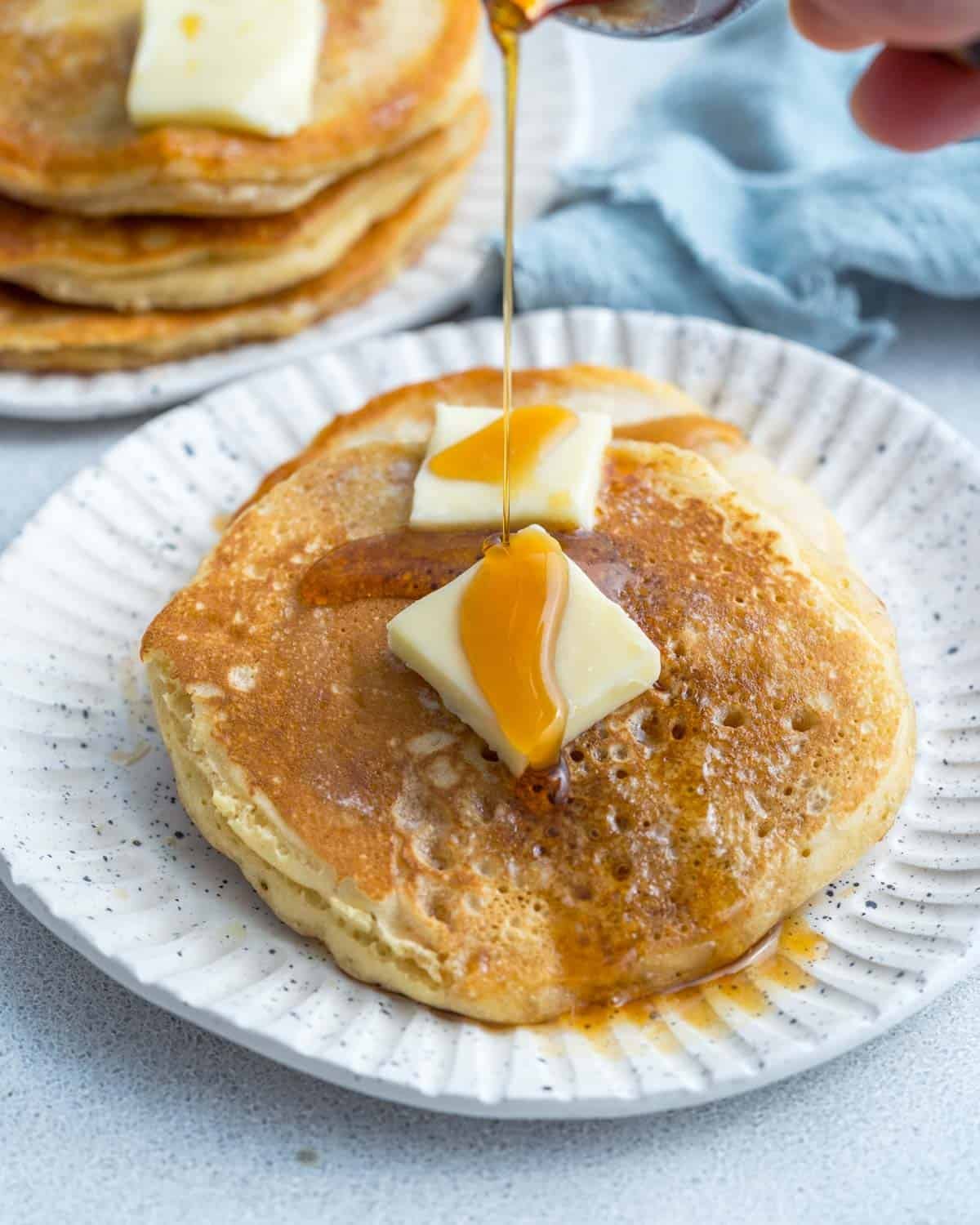 Cooked pancakes on a white plate with pats of butter and a drizzle of maple syrup.