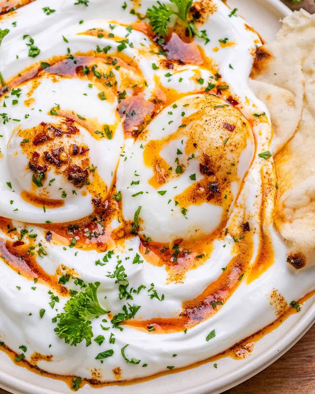 Turkish eggs in a white bowl topped with parsley and a naan.