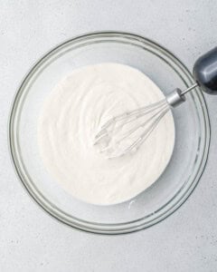 Clear glass bowl showing the mixture of yogurt, protein powder, and honey being whisked with a metal hand whisk until smooth and creamy.