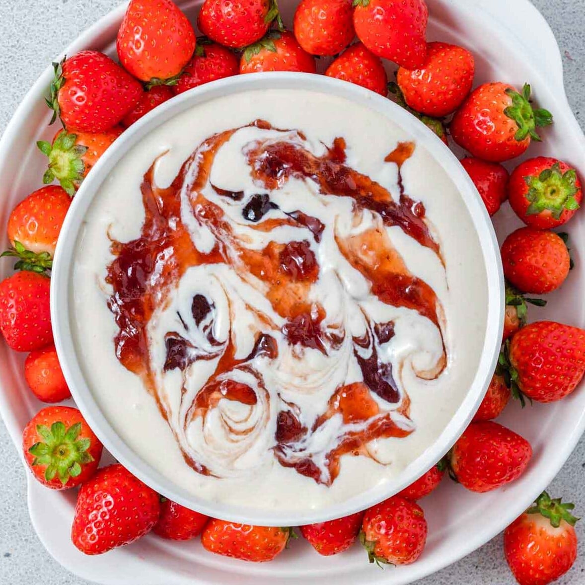 Close up of finished strawberry cheesecake dip in a white bowl with strawberry preserves swirled throughout, and sliced strawberries on top, surrounded by fresh strawberries on a white plate.