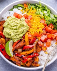 A closeup of a white bowl with chicken, peppers, onions, lettuce, rice and cheese.