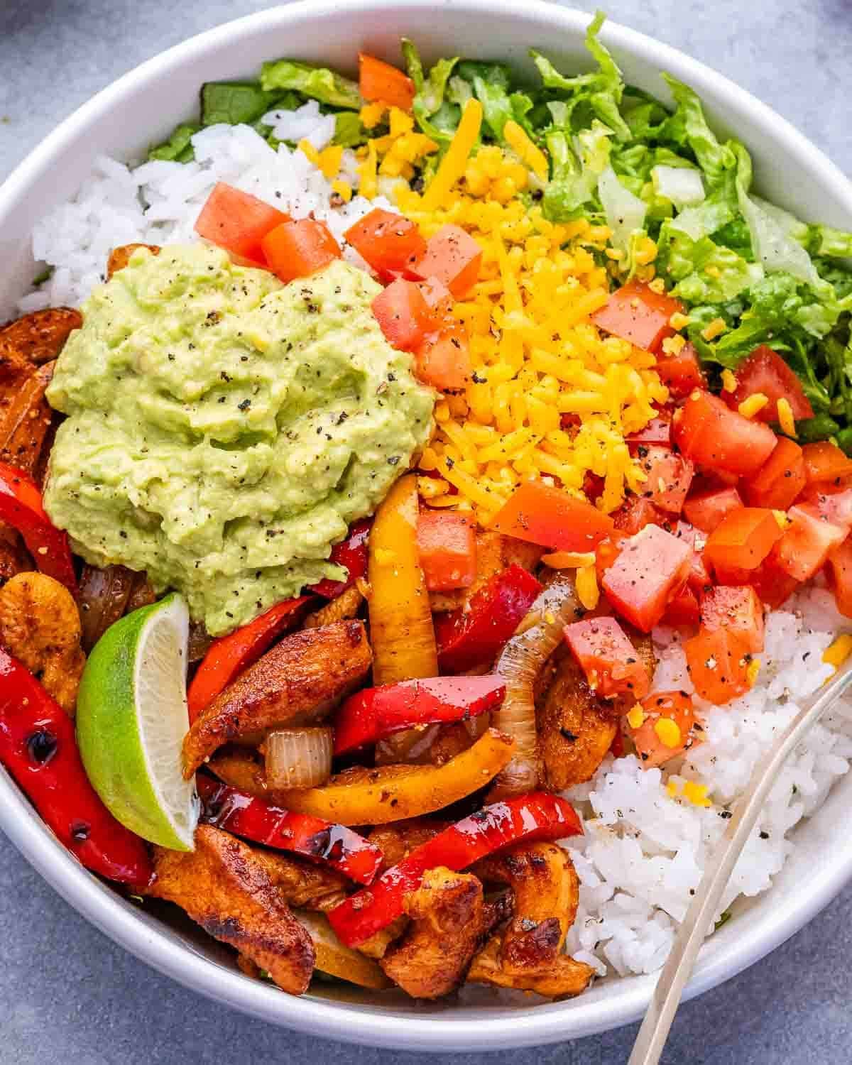 A closeup of a white bowl with chicken, peppers, onions, lettuce, rice and cheese.