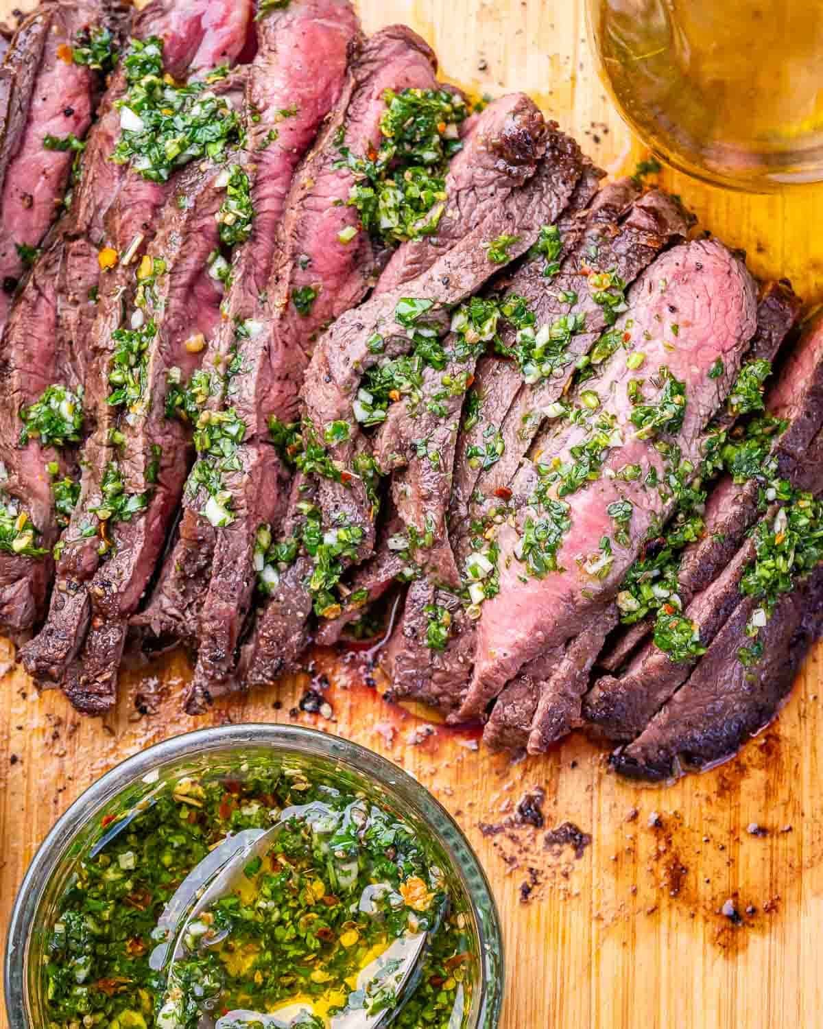 Sliced grilled steak on a wooden board with homemade chimichurri.