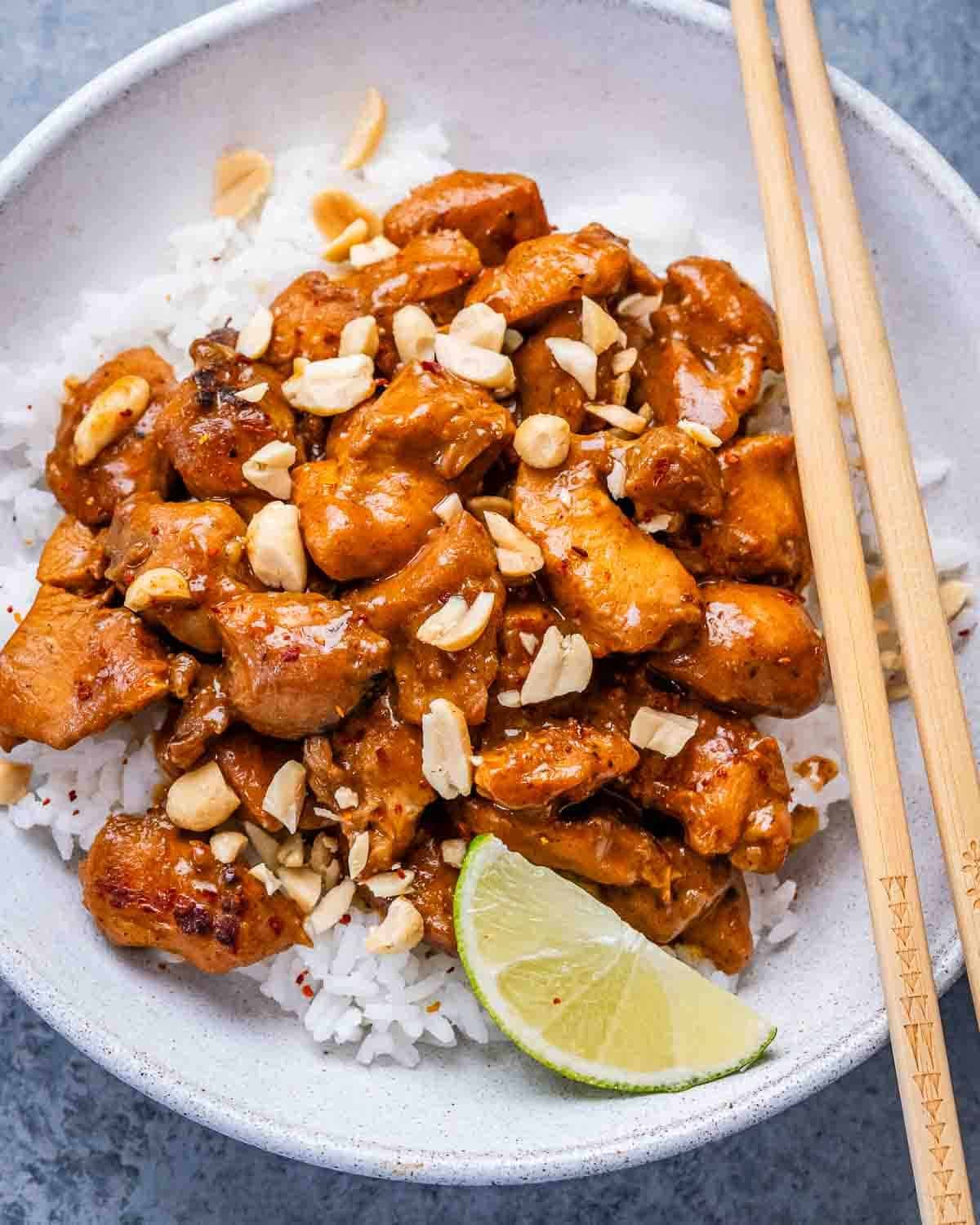 A white bowl and chopsticks with rice, a lime wedge, and peanut butter chicken.