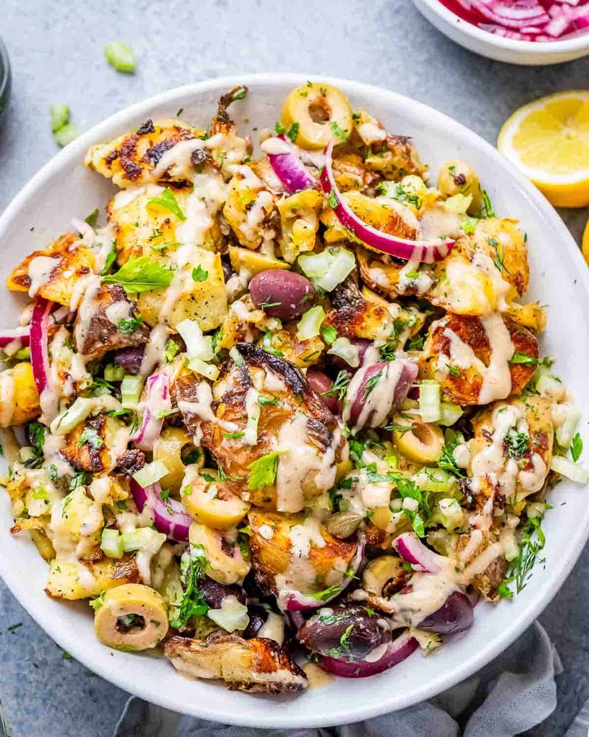 smashed potato salad with veggies and olives in a deep white dish.