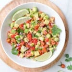 round bowl filled with avocado salsa with lime wedges on them.