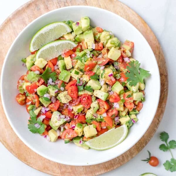 round bowl filled with avocado salsa with lime wedges on them.