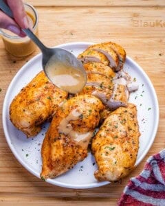 A spoon pouring onion gravy over cooked chicken breasts on a white plate.