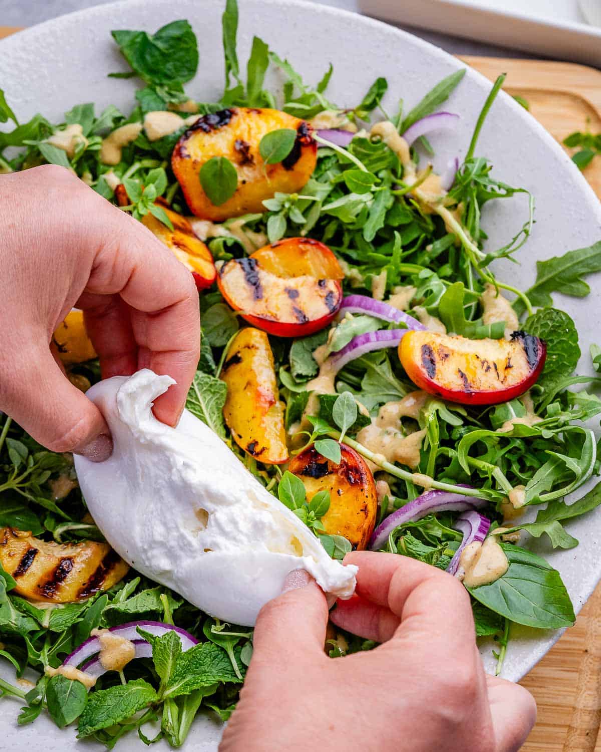 Hands tearing burrata over a peach salad.