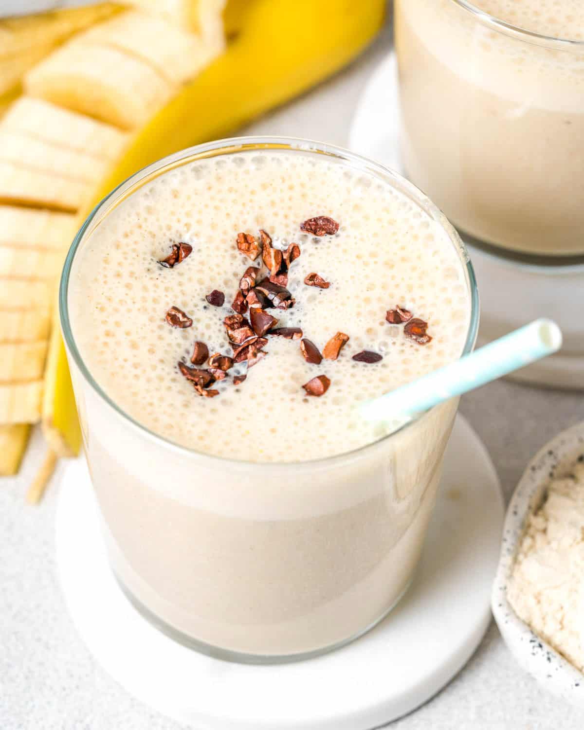 a glass filled with banana protein shake topped with cocoa nibs and a straw in the cup.
