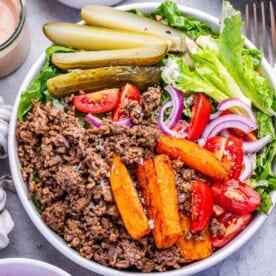 a bowl with cheeburger ground beef and lettuce topped with sweet potato fries, tomatoes, and sliced pickles.
