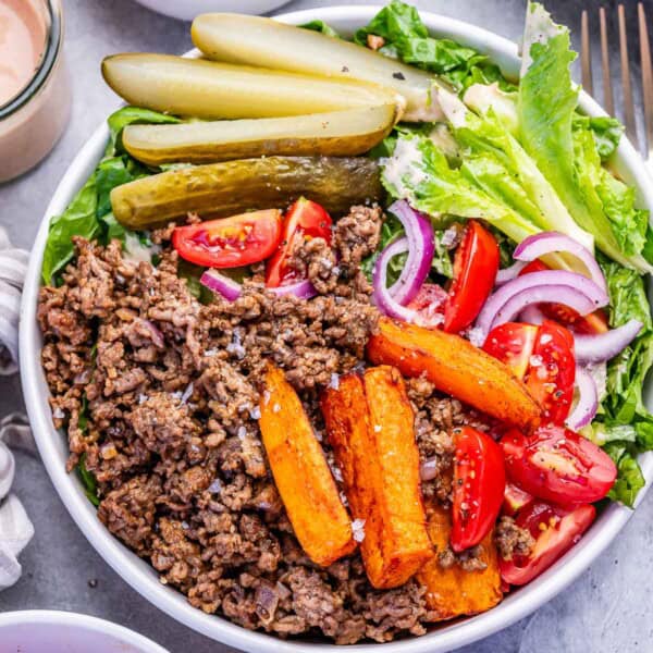 a bowl with cheeburger ground beef and lettuce topped with sweet potato fries, tomatoes, and sliced pickles.