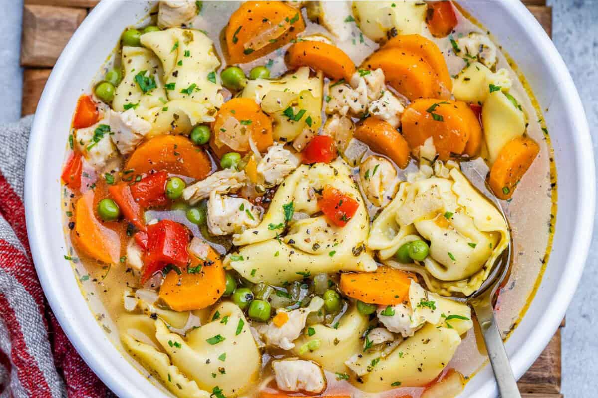 A close-up of a bowl of chicken tortellini soup with carrots, peas, and bell pepper.