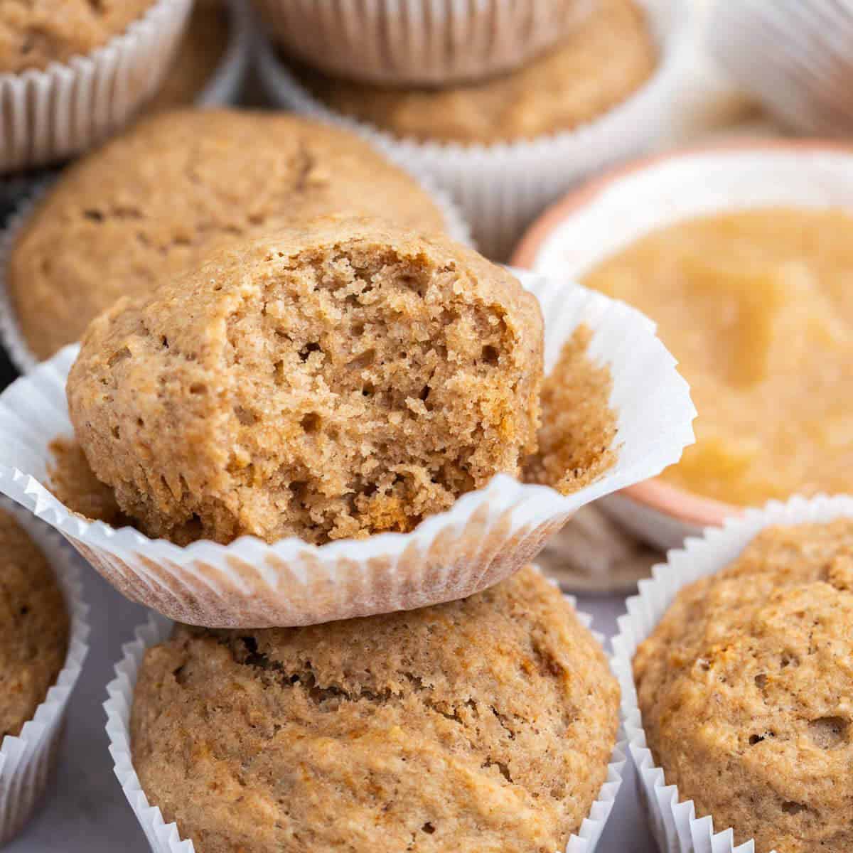 An applesauce muffin with a bite taken out, surrounded by more muffins and a bowl of applesauce.