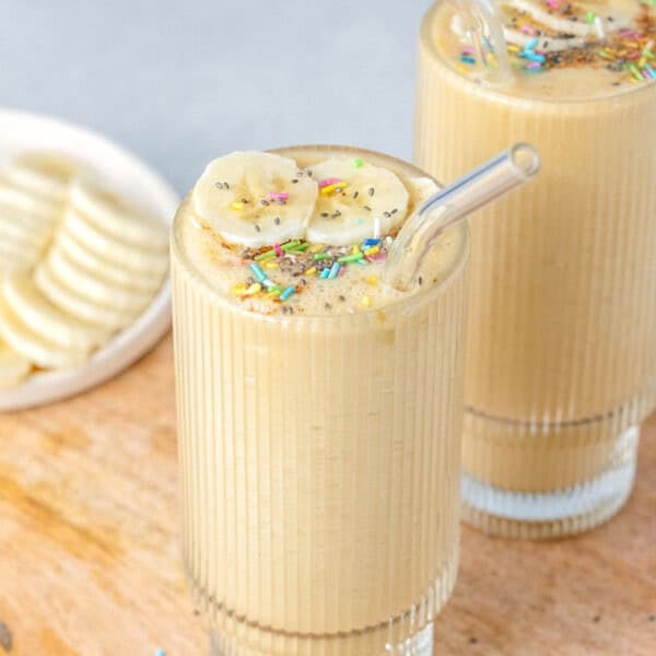 a tall glass filled with vanilla protein shake topped with sprinkles and sliced bananas.