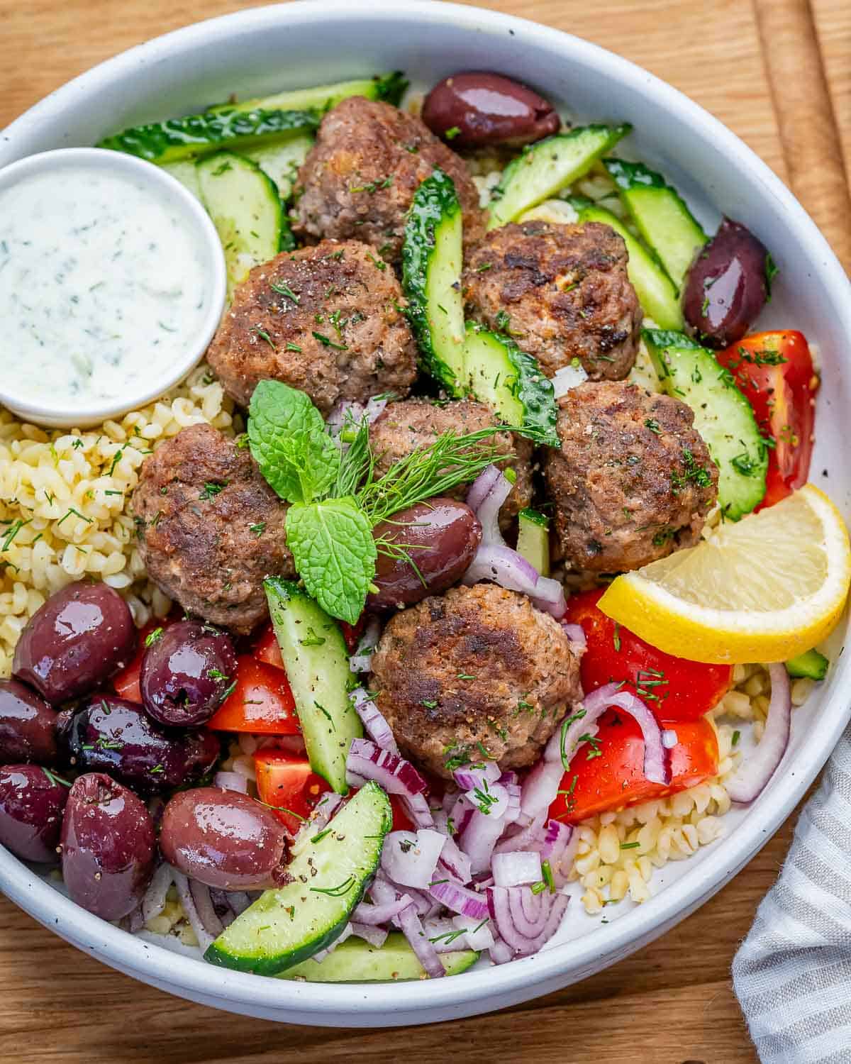 greek balls assembled in a bowl with cooked bulgur topped with kalamata olives, cucumber, and onions.