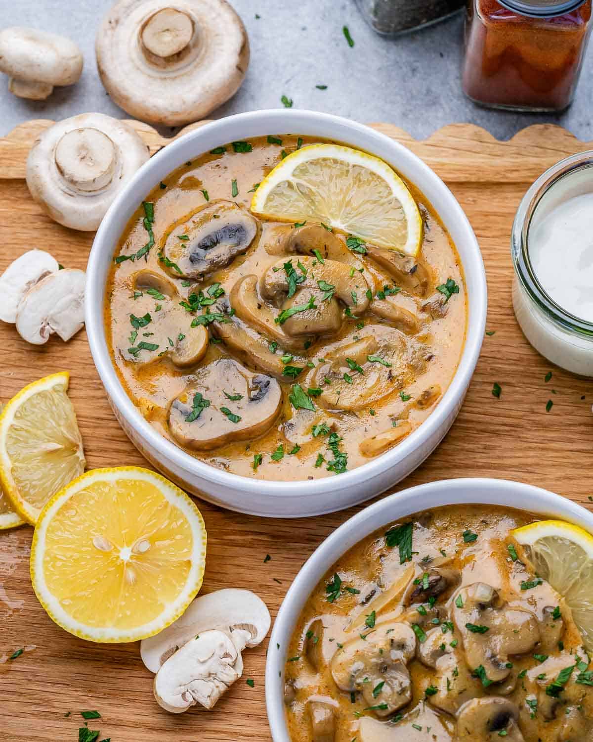 Two white bowls of Hungarian mushroom soup on a wooden board, with a wedge of lemon.