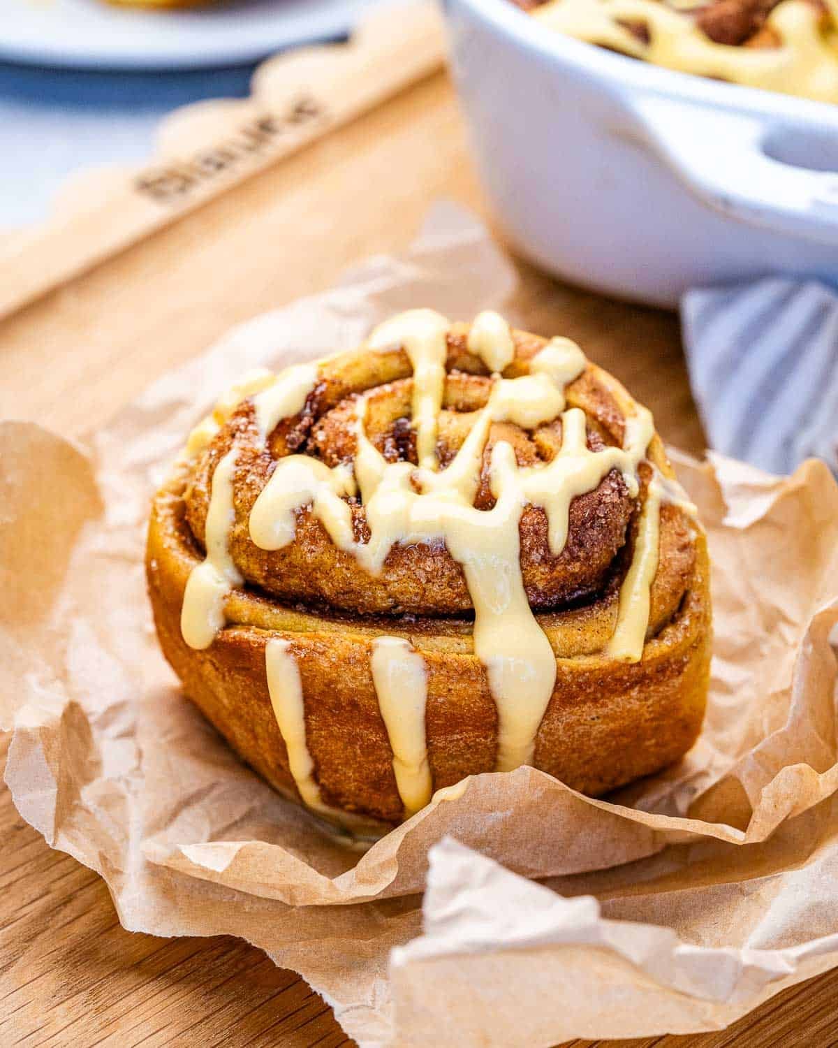 A pumpkin cinnamon roll drizzled with glaze, on a piece of parchment paper on a wooden board.