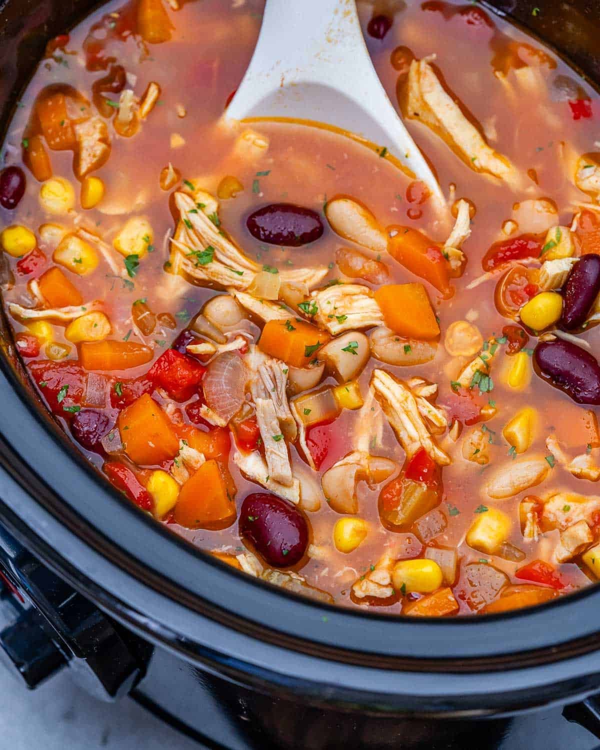 A slow cooker filled with chicken tortilla soup with corn and beans and a spoon.
