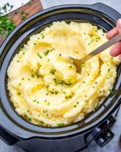 A spoon scooping up slow cooker mashed potatoes topped with chives.