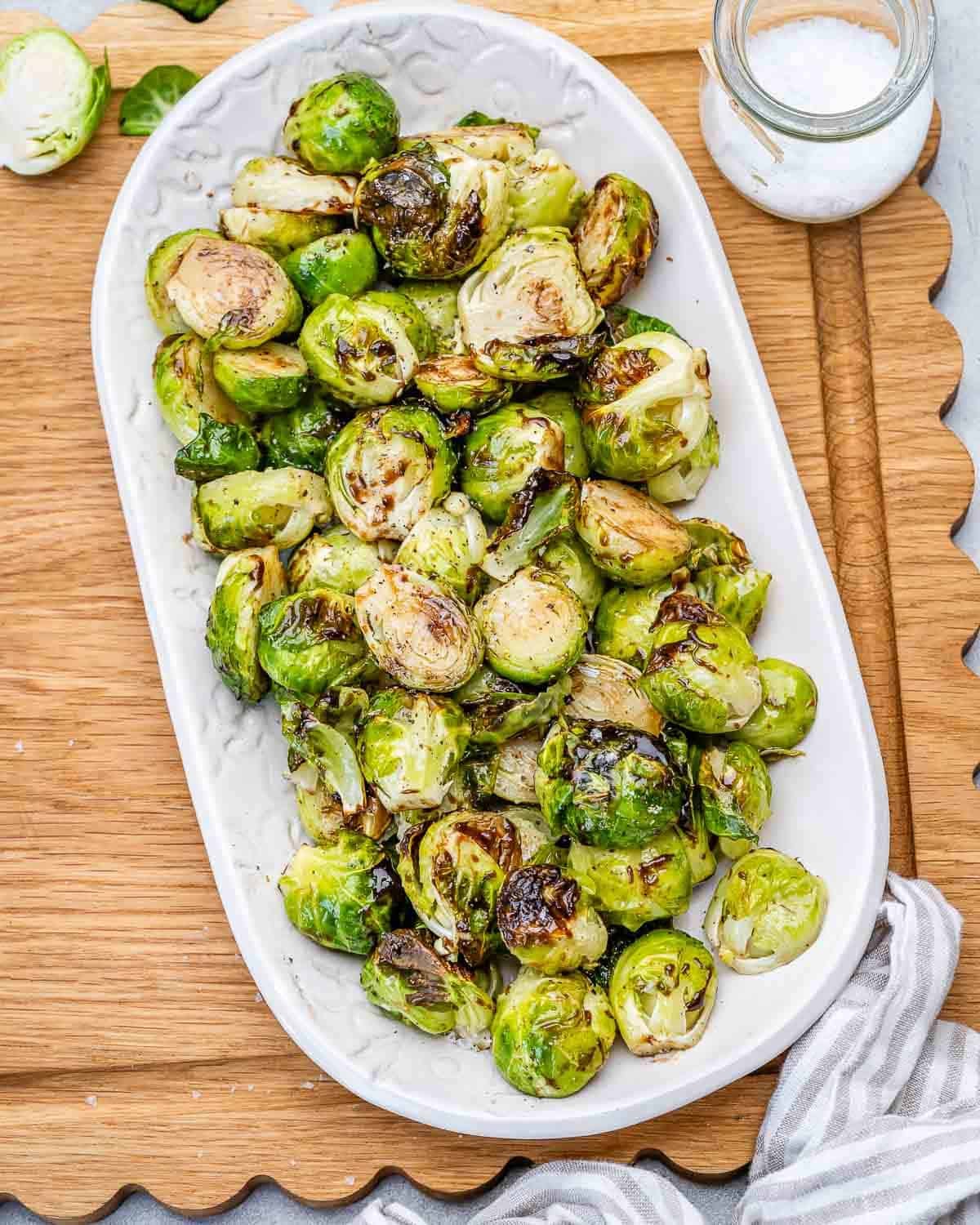 A white oval plate of roasted brussels sprouts with a balsamic glaze, on a wooden board.