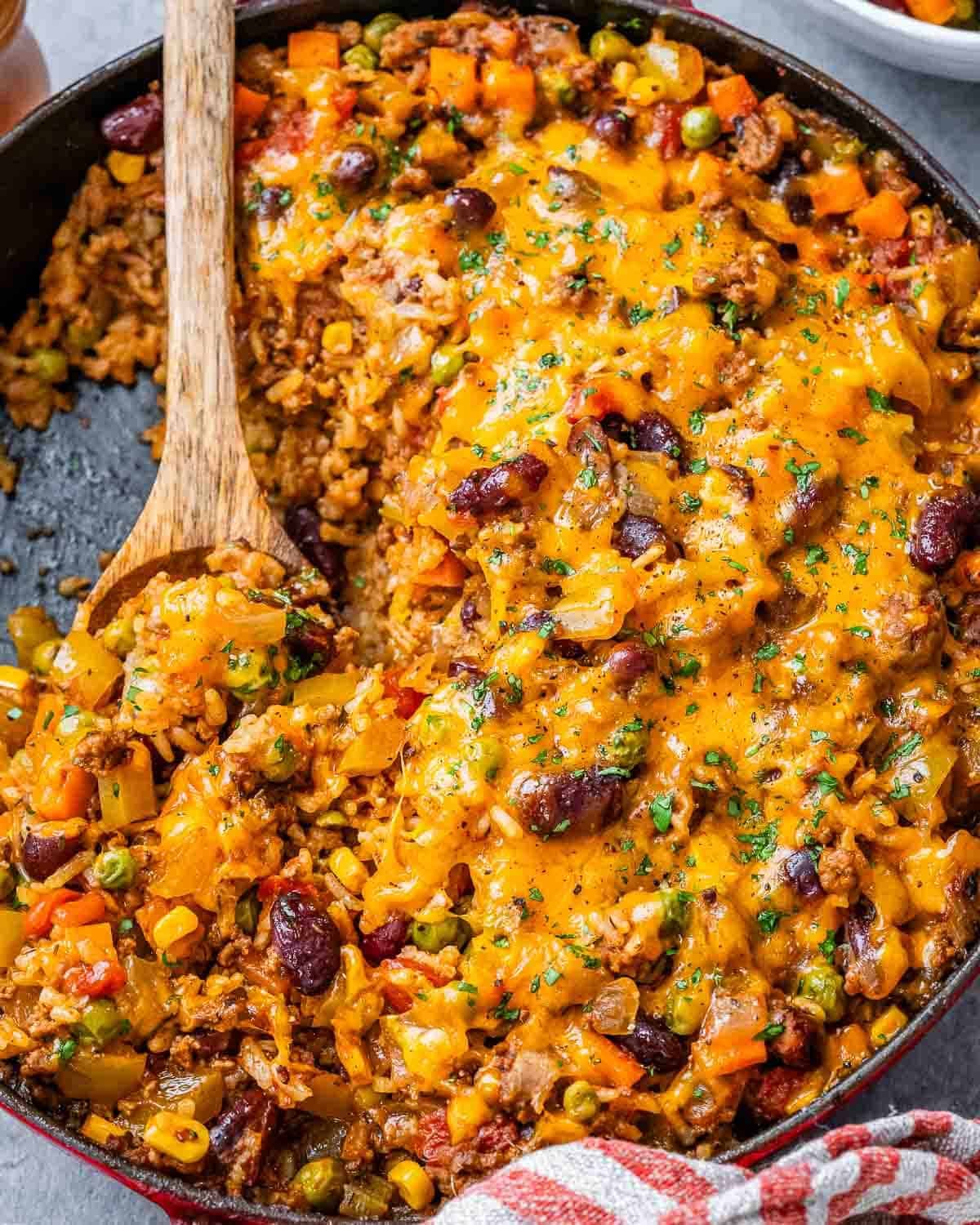 A skillet of ground beef and rice casserole with a wooden spoon scooping some out.